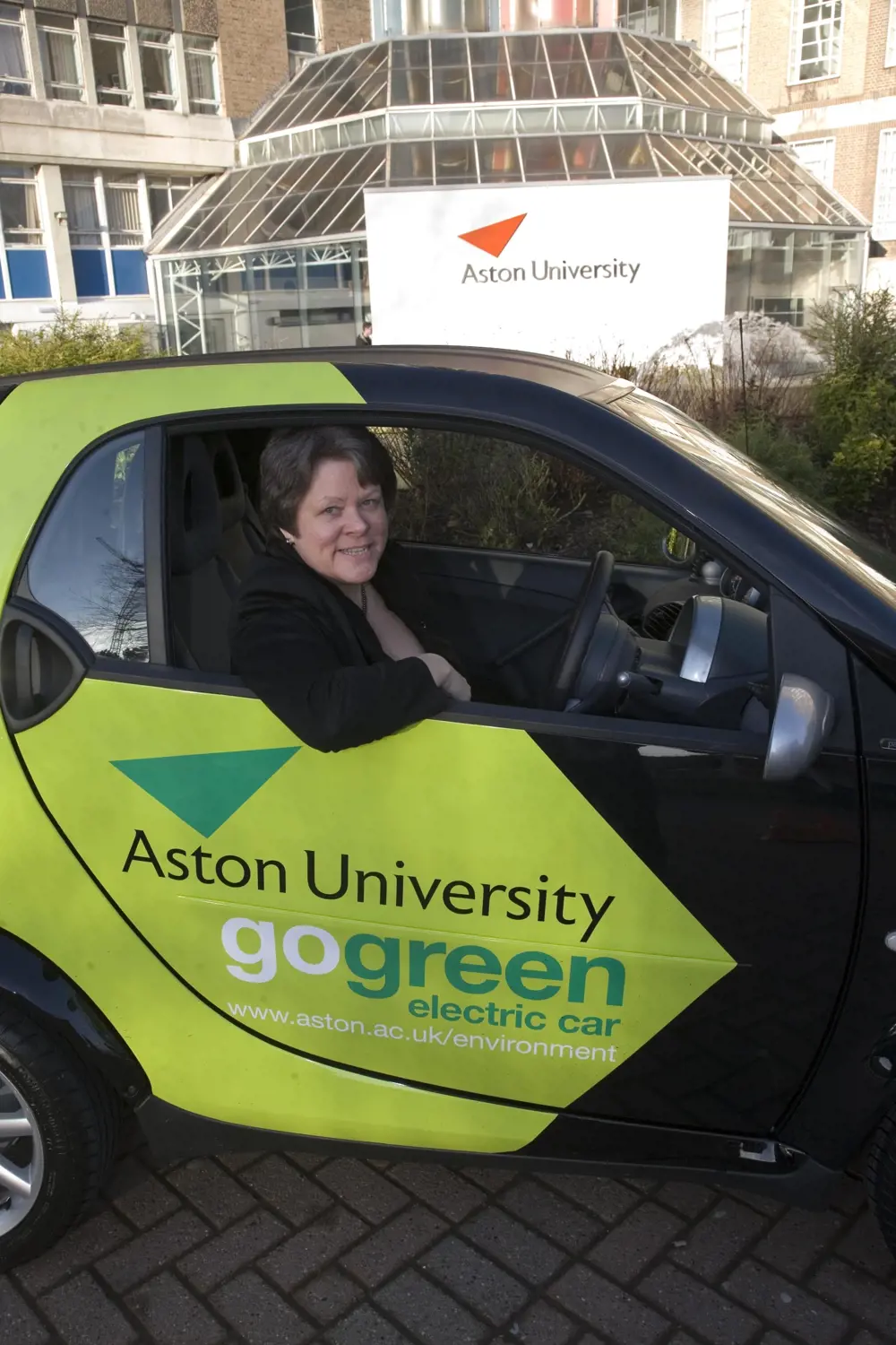 Baroness Brown inside a SMART electric car, that has advertising for 'Aston University' and 'go green'.