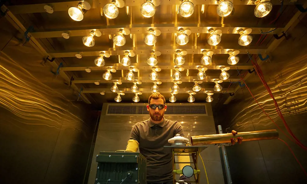 A male engineer standing behind a desk with scientific equipment, wearing goggles and gloves, working in a room that is lit up with bright bulbs on the ceiling.