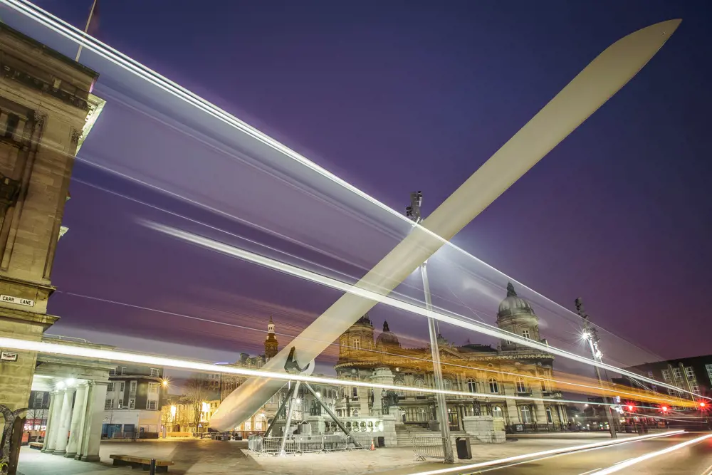 A long-exposure photograph of a white turbine blade at night supported to sit diagonally with light trails created by buses streaming across the photograph.