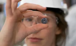 A close-up of a scientist wearing goggles and a safety hat holding a small metal round object. 