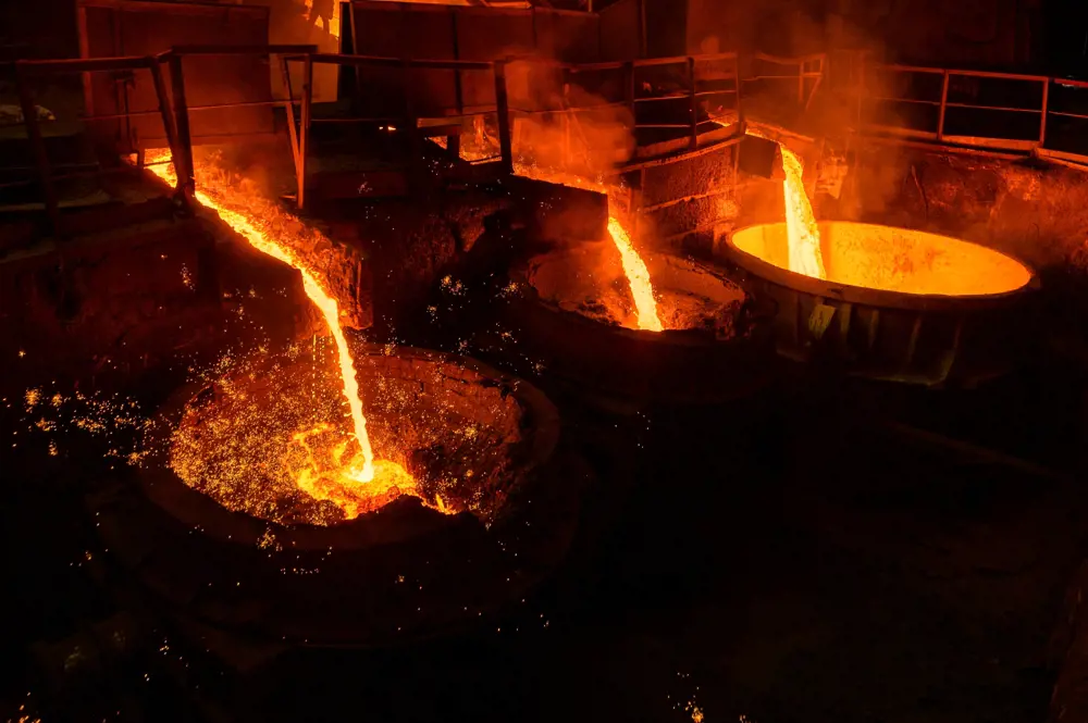 Streams of molten metal and slag, glowing orange, are pouring into three vessels from the base of different furnaces in a steelworks.