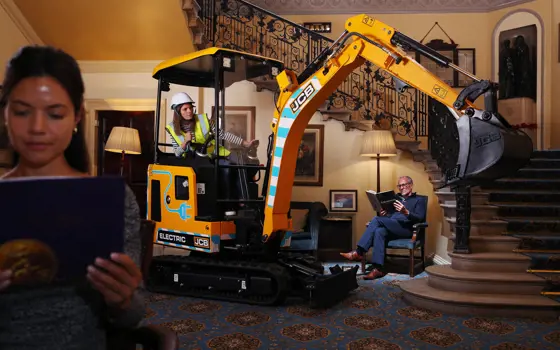 An electric JCB placed inside of a house, with two people reading books around it to demonstrate it's quietness. 