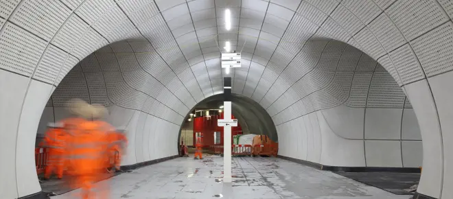 The inside of the underground Elizabeth Line station while it was under construction with glass fibre reinforced concrete cladding panels lining the walls. A blurry worker in construction gear is walking through the tunnel. 