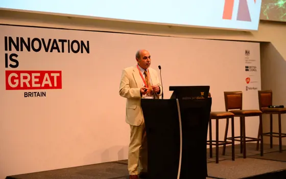 Professor Sir Saeed Zahedi OBE FREng standing on a stage in front of a microphone, giving a talk, with a board saying 'Innovation is great Britain' behind him.