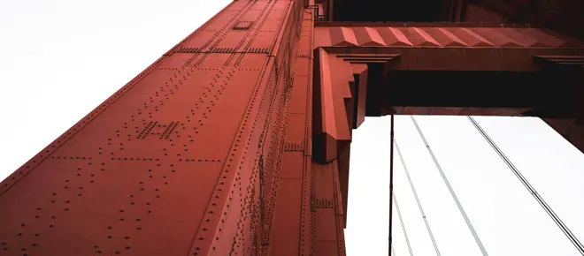 A worms-eye view of a red steel tower of the Golden Gate Bridge on an overcast day.