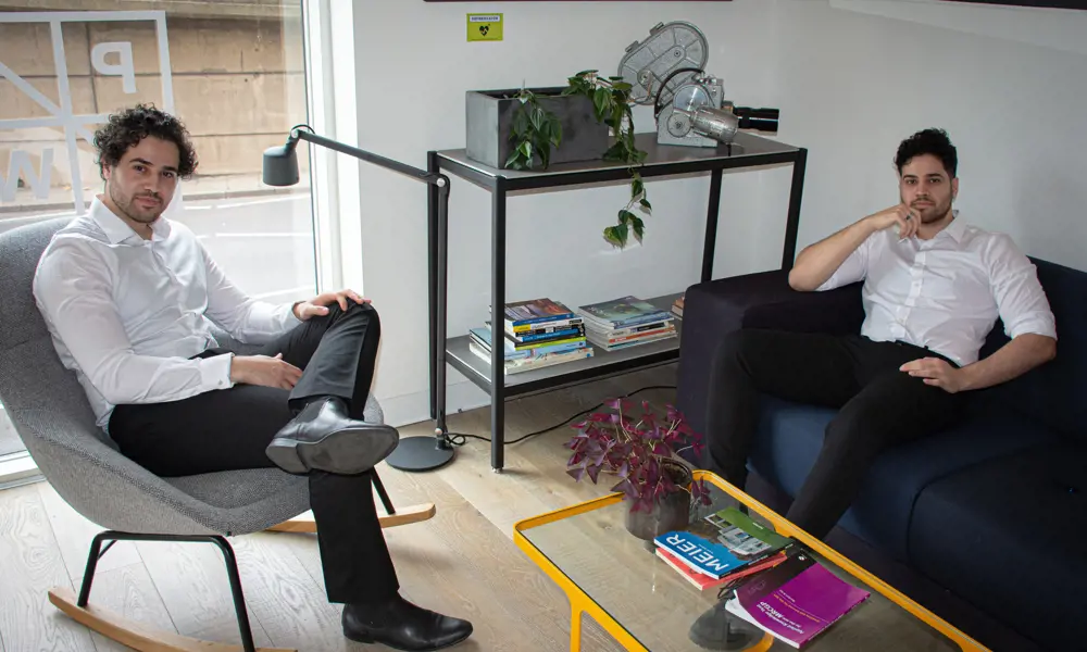 A photograph of Hussan sitting on a grey chair in the left corner and Hussain sitting on a sofa in the right corner of the photograph. Both are looking at the camera with relaxed postures.