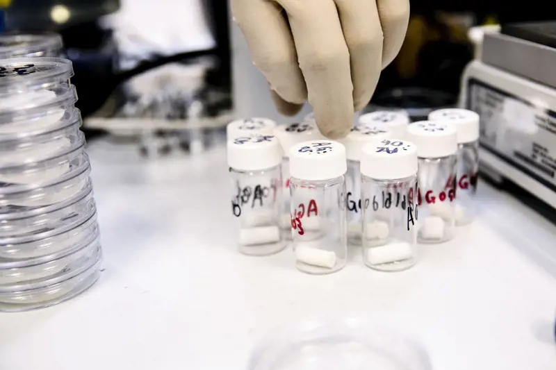 A hand with gloves on reaching for small containers that contain a white tablet-shaped material inside.