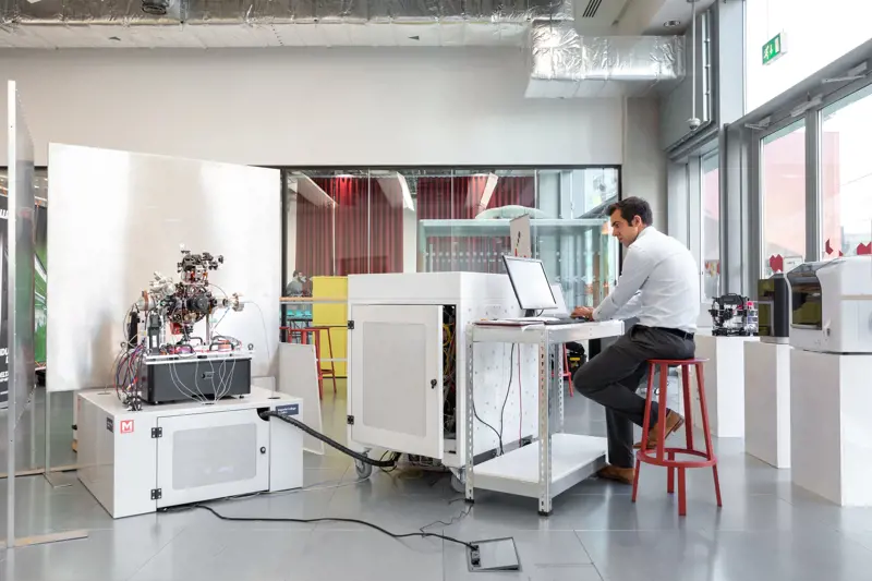 A person sitting on a stool looking at a computer which is connected to a quantum accelerometer in a lab.