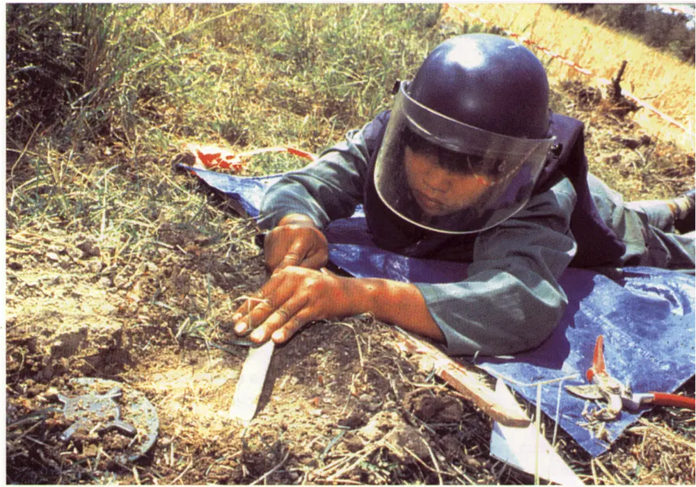 A person lying on the ground in front of a landmine to try and demine it, wearing a protective face shield and vest. 