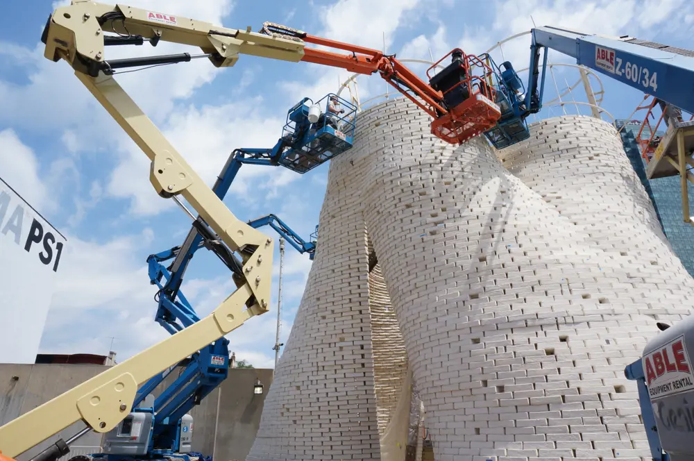 People on two cherry pickers in front of a tower made from white mycelium bricks. They are finishing the tower.