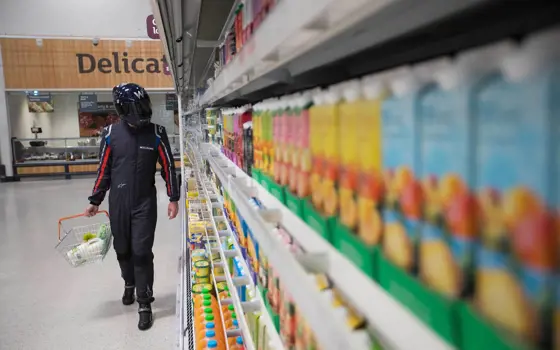 A person in a williams' racing suit and helmet in a supermarket, holding a basket of groceries.