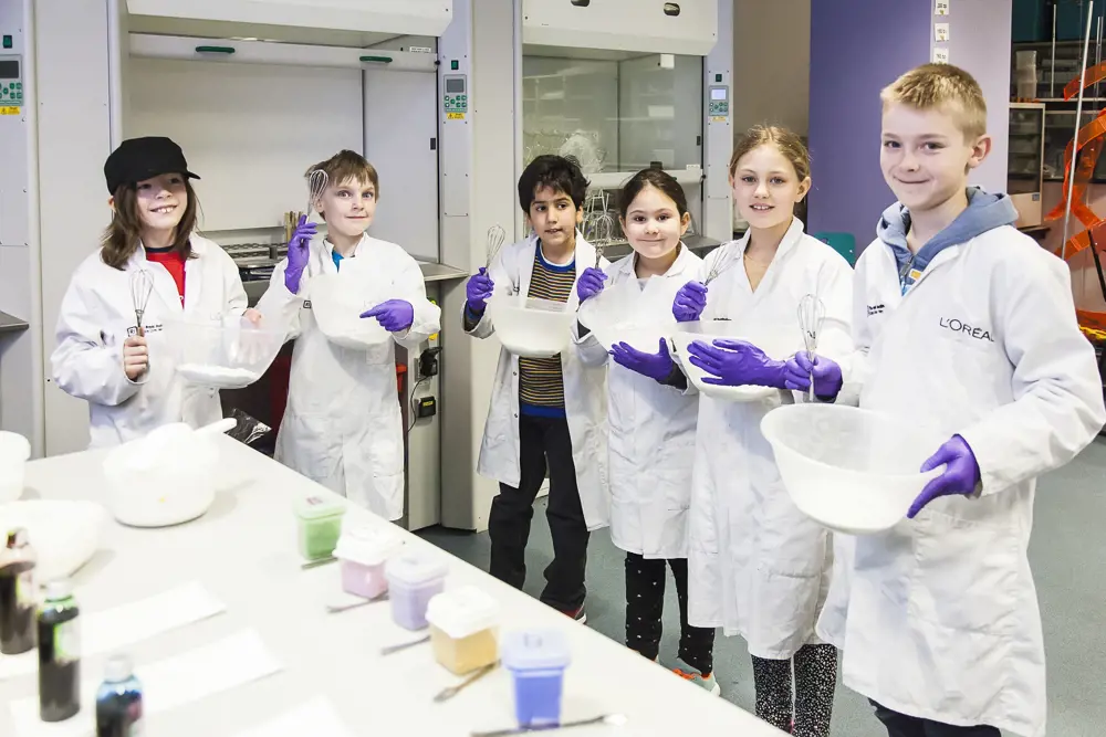 Six young people in a laboratory, wearing lab coats and holding mixing bowls and whisks. There is a table in front of them with different coloured substances in containers. 