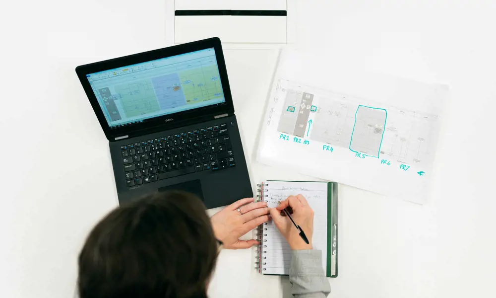 An aerial view of an engineer writing in an A5 notebook, with a laptop and paper highlighted on the desk in front of them.