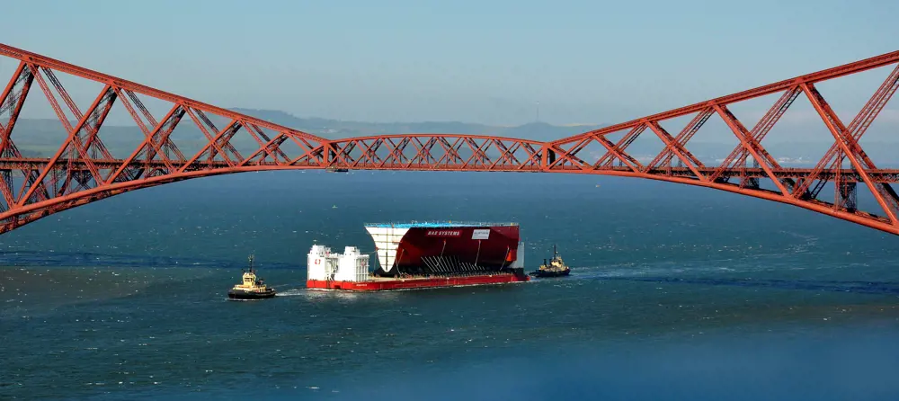 A large BAE Systems barge under a bridge in the ocean.