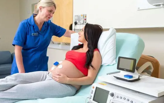 A pregnant women lying down on a hospital bed, talking to a nurse. The women is wearing the Monica Novii wireless patch system on her belly