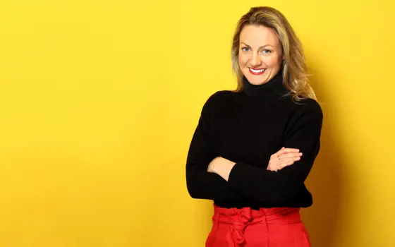 A headshot of Sinead O’Sullivan smiling at the camera, with her arms crossed and a yellow background.