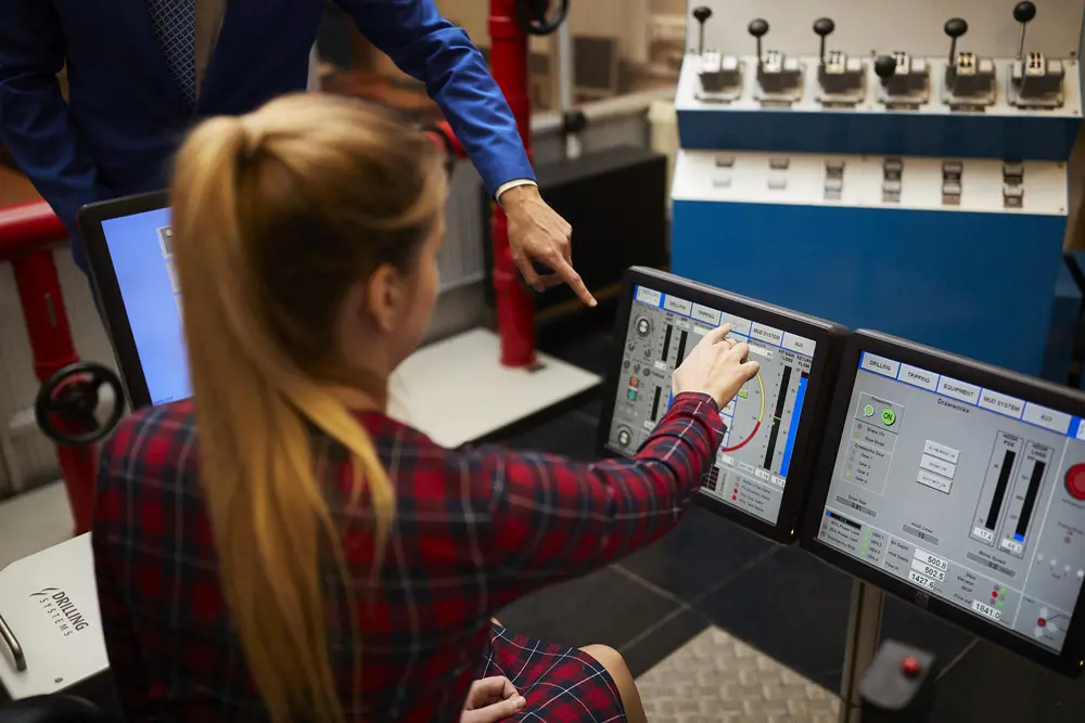 The back of a woman with long blonde hair in a ponytail wearing a read dress. She is pointing at data across several screens and someone unseen from the side also points at the screen