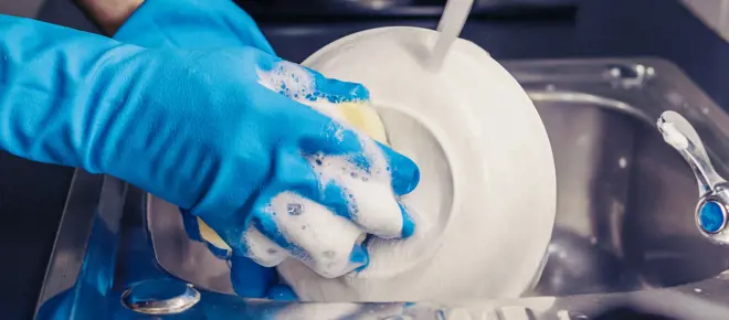 Someone's hands wearing blue washing up gloves, cleaning a plate with a sponge and washing up liquid in the sink