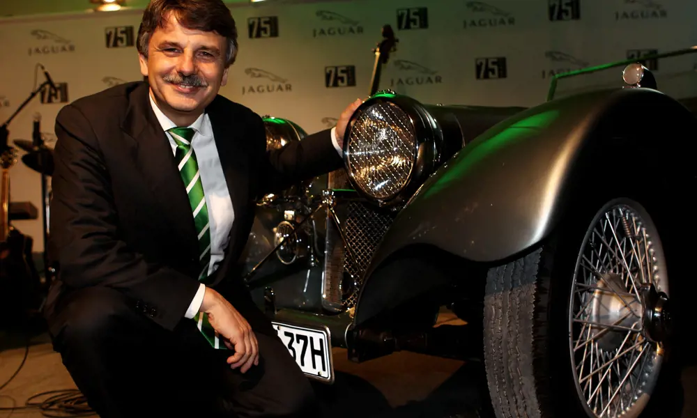 Ralf Speth, crouching next to an old Jaguar at a formal event in Germany.