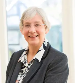 A smiling white woman with glasses and short hair, wearing a grey blazer, looks at the camera