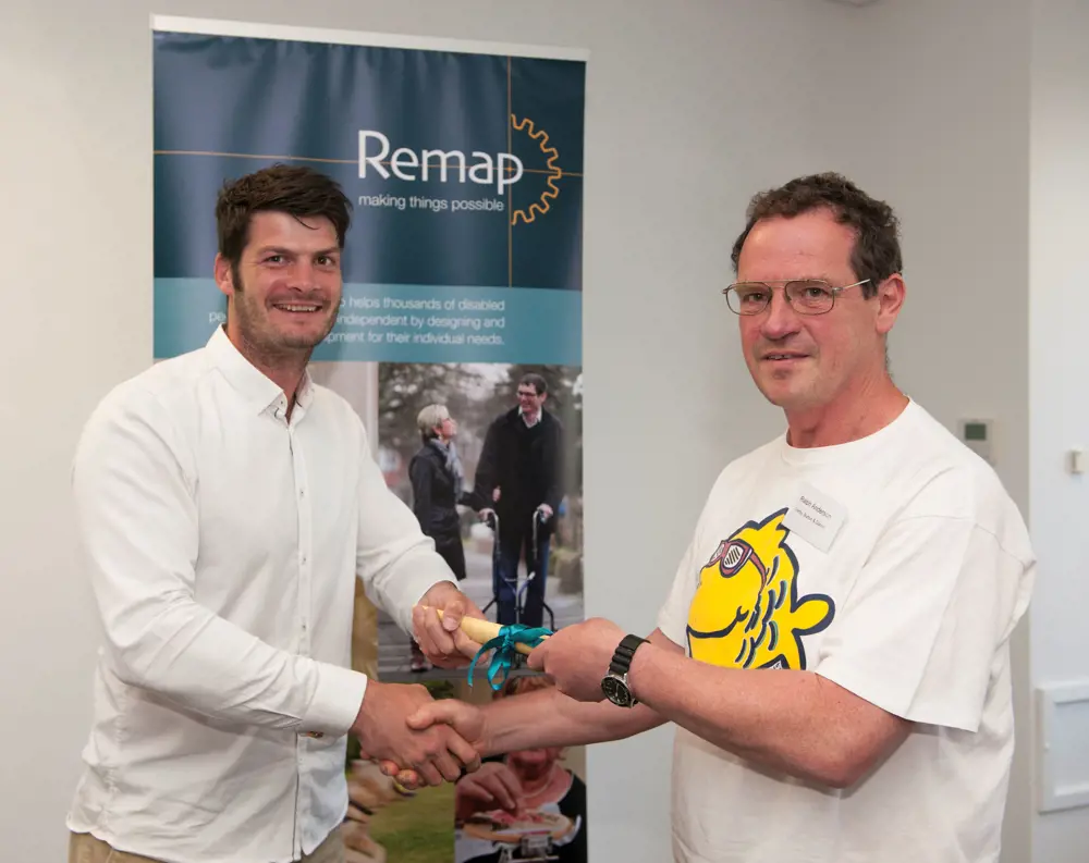 Dave Henderson MBE shaking hands with Ralph Anderson and giving him an award that is rolled up. Both are smiling at the camera. 