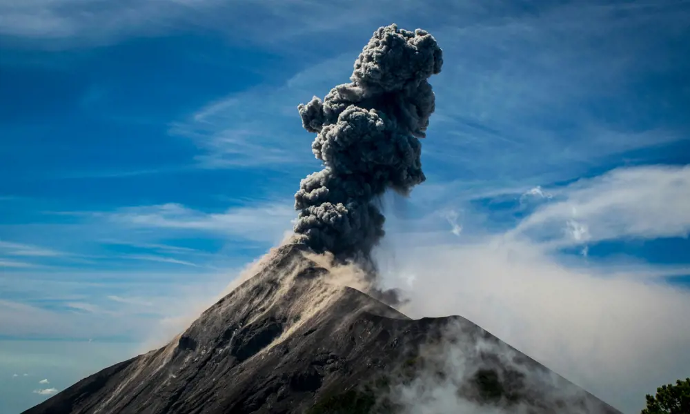 A volcano with a plume of dark grey smoke and ash emerging from the top, surrounded by misty clouds and with a bold blue sky behind.