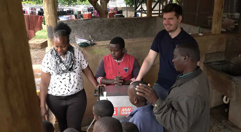 A group of adults and children gather around a portable washing machine in an outdoor sheltered area, with two people standing behind the machine and several children seated in front.