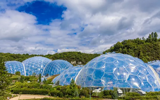 Large transparent geodesic domes of the Eden Project sit among green hills under a bright, partly cloudy sky.