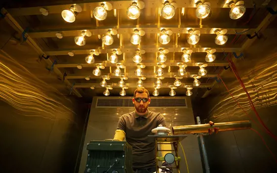 A male engineer standing behind a desk with scientific equipment, wearing goggles and gloves, working in a room that is lit up with bright bulbs on the ceiling.