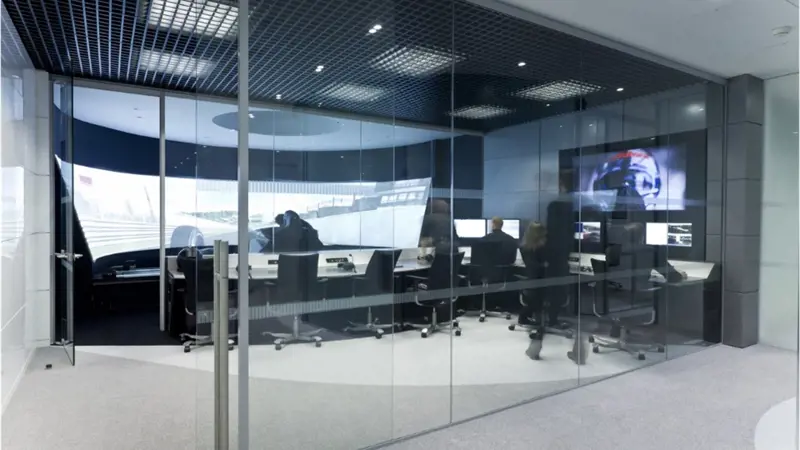 A control room with 8 swivel chairs arranged around desks facing a huge wraparound screen displaying the simulated view from a racecar.