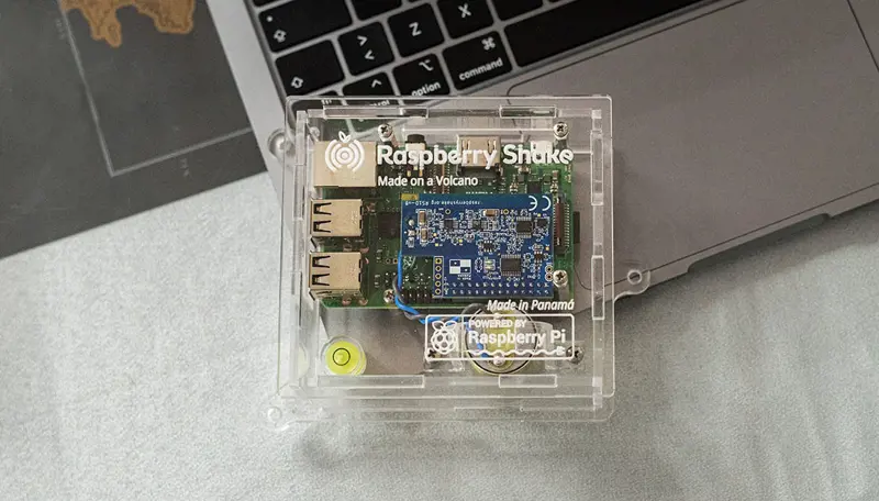 A small computer board sits within a clear Perspex box on the edge of a laptop. The text on the box reads: Raspberry Shake. Made on a volcano