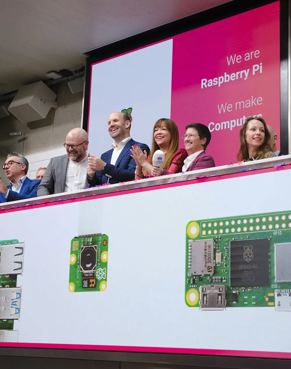 A group of six people - three men and three women - stand on a balcony. The balcony has images of Raspberry Pi computer and a pink and white poster behind the people reads: We are Raspberry Pi. We make computers.s