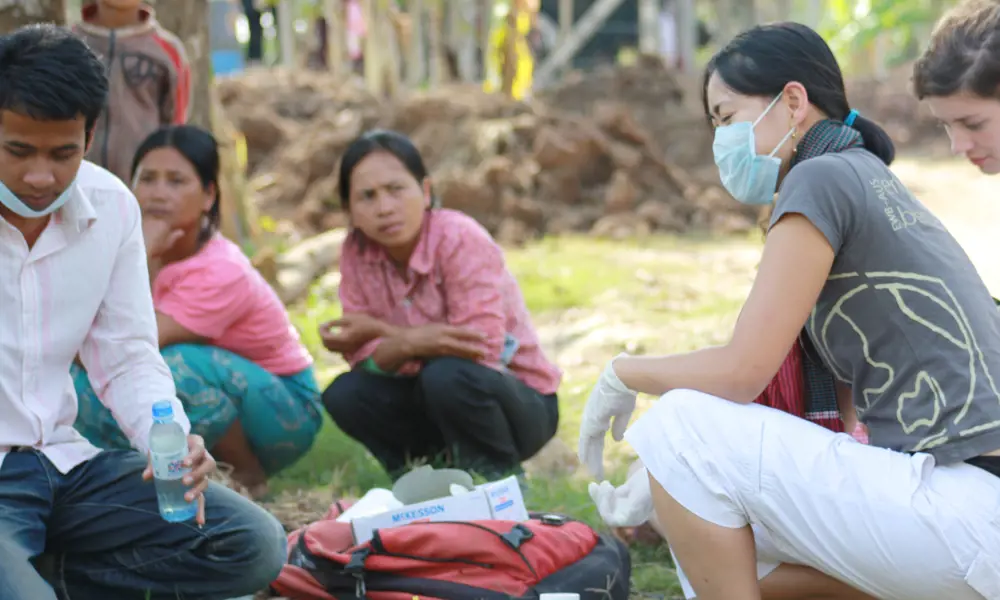 Dr Anh Tran crouching down with a mask and gloves on, with chemical bottles in front of her on the ground, being watched by local people in Cambodia.
