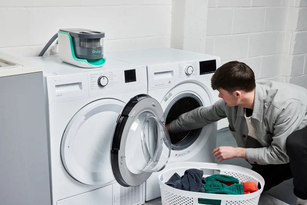 A man with dark hair loads washing into a machine from a white basket full of clothes next to him. There is a filter attached to the top of the machine
