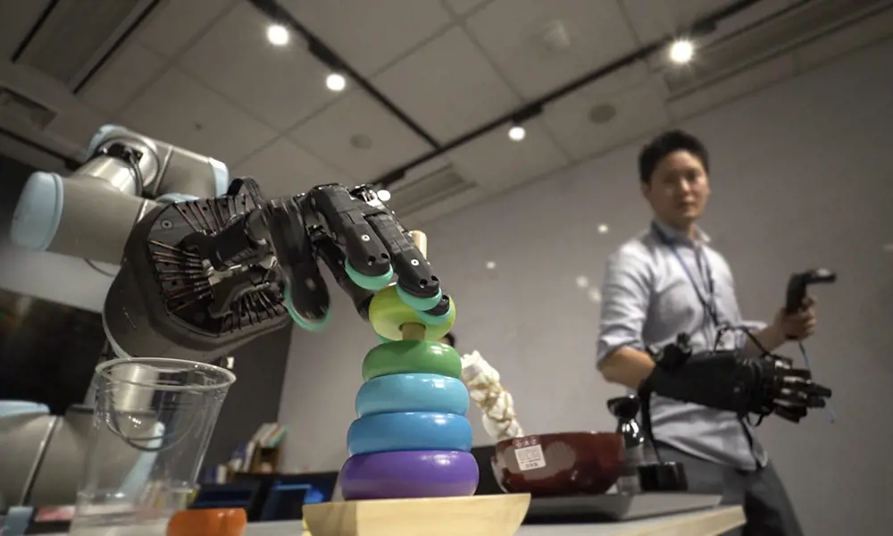 The Shadow Hand robot arm sliding a ring off a wooden stick. The hand is being controlled by a person in the background wearing a large glove. 