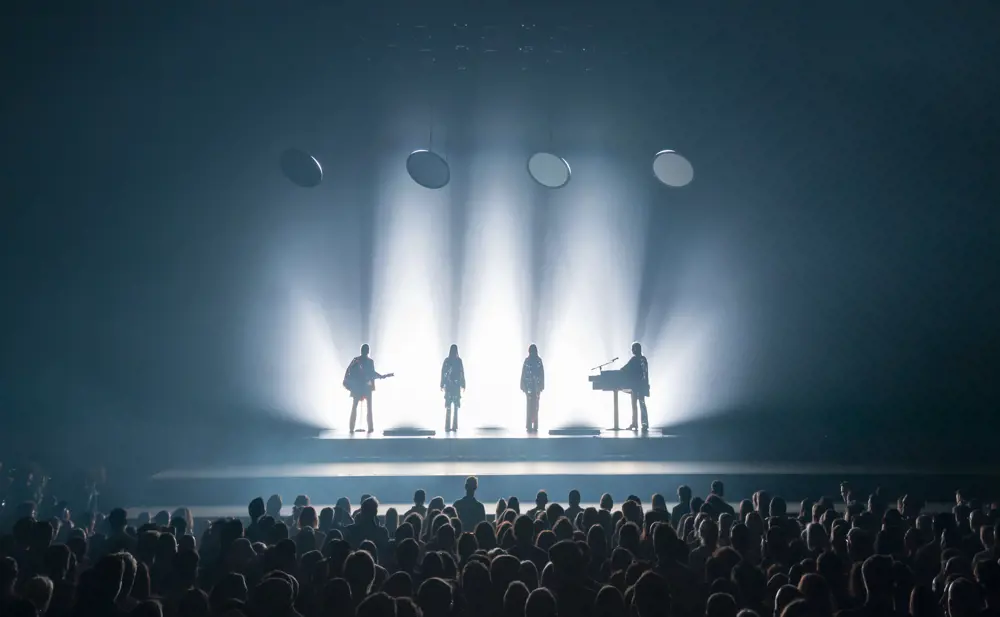 Two singers and two musicians playing a guitar and keyboard are brightly backlit by spotlights on a stage in front of a large crowd.