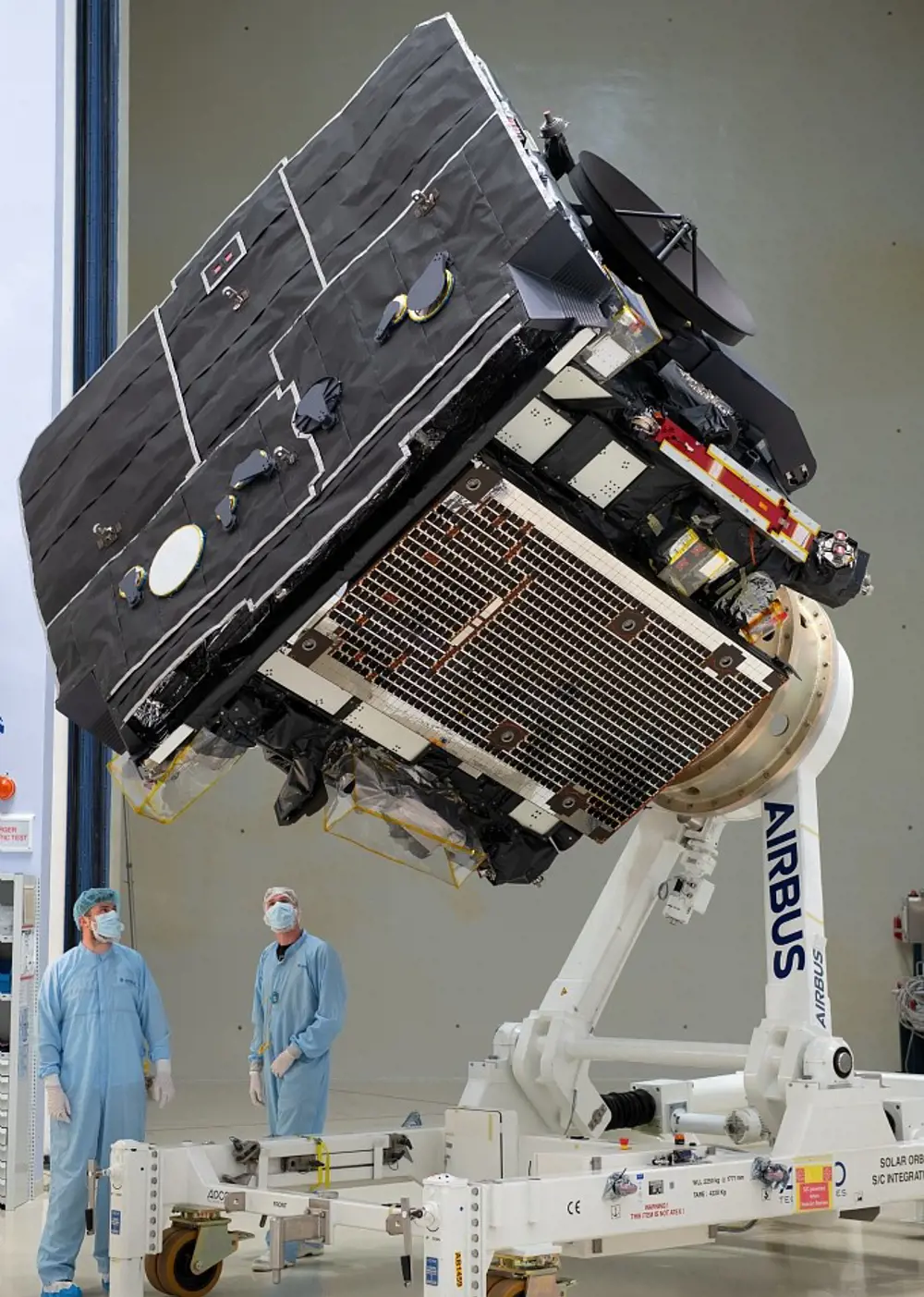 The solar orbiter at IABG test facilities in Germany, being packaged and transported. Two people in masks and protective suits are shown looking up at the orbiter for scale.