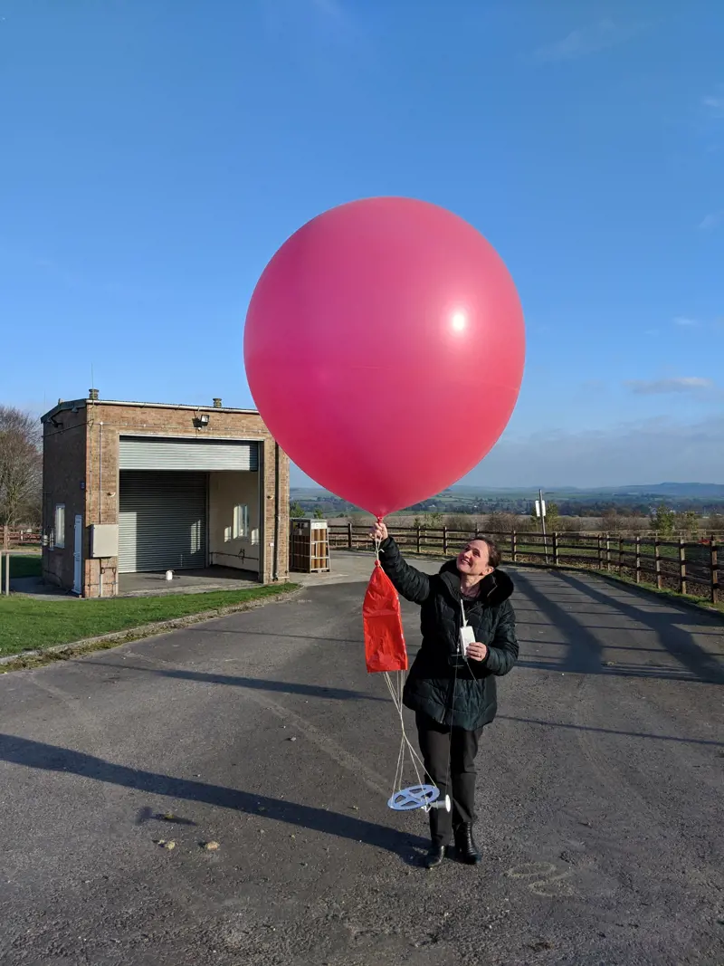 Penny wearing a black coat, holding a large red weather balloon with blue skies and green countryside behind her.