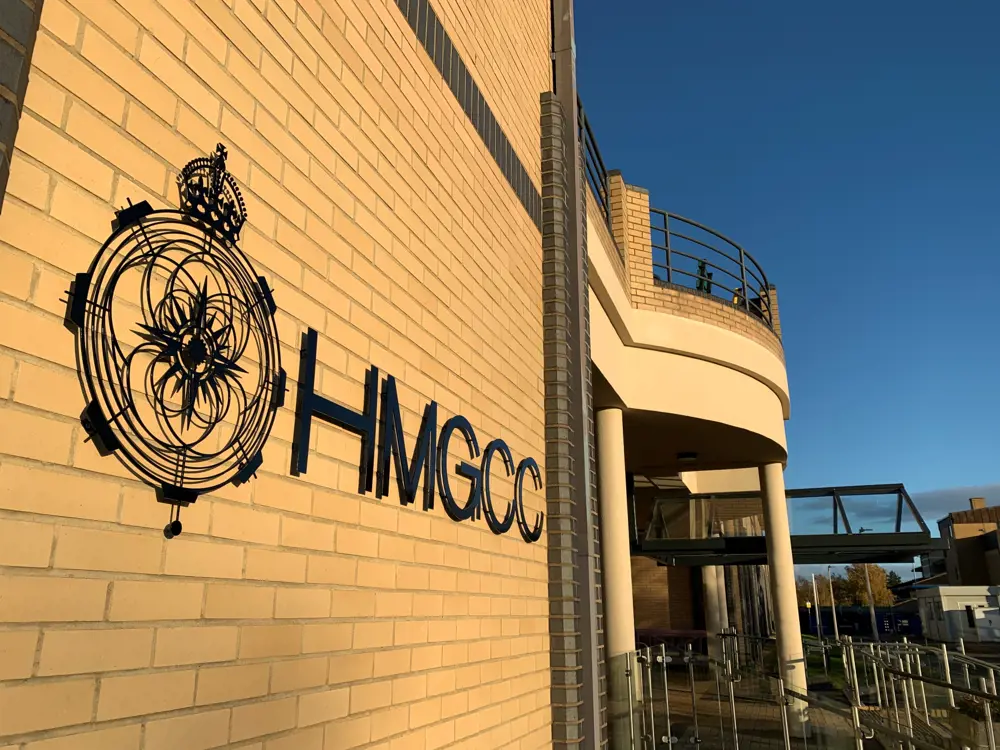 The facade of a government building with the wording "HMGCC" affixed to the wall, as the day approaches sunset.