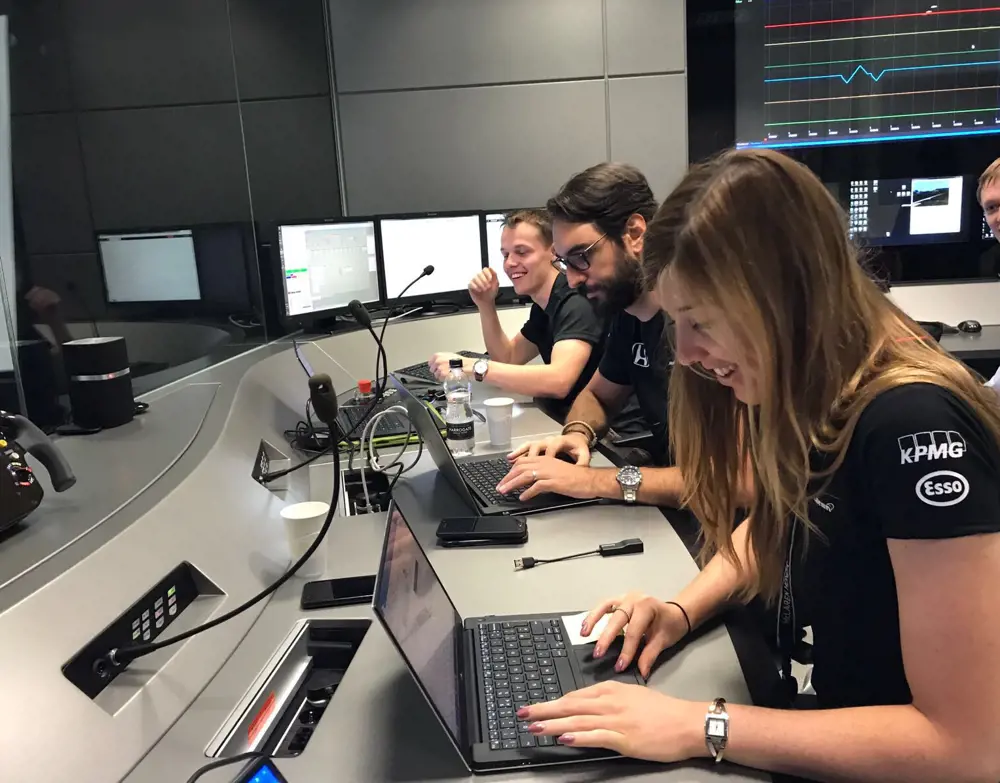 Three people - one woman and two men - sit along a desk working on computers. They have microphones in front of them.