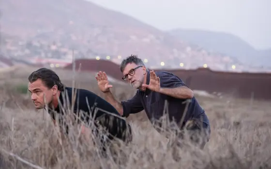 Leonardo DiCaprio and Paul Thomas Anderson are crouching in tall dry grass on a hillside, with Paul gesturing as if framing a shot, and a blurred view of mountains and a distant town in the background.