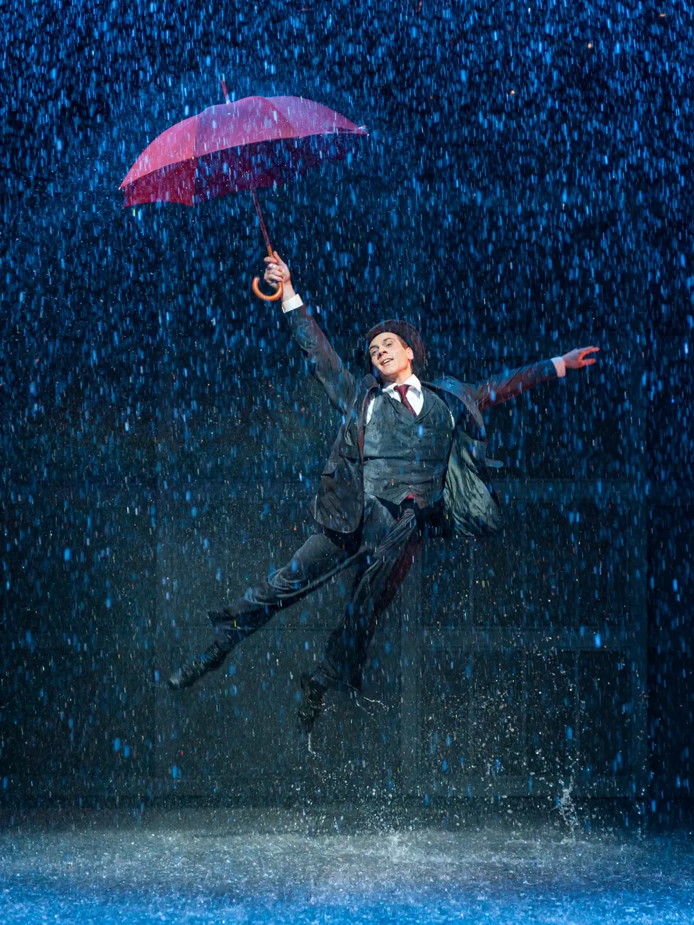 A performer on stage, jumping in the air while water is falling down around them and they are holding an open umbrella and pointing it towards the ceiling.