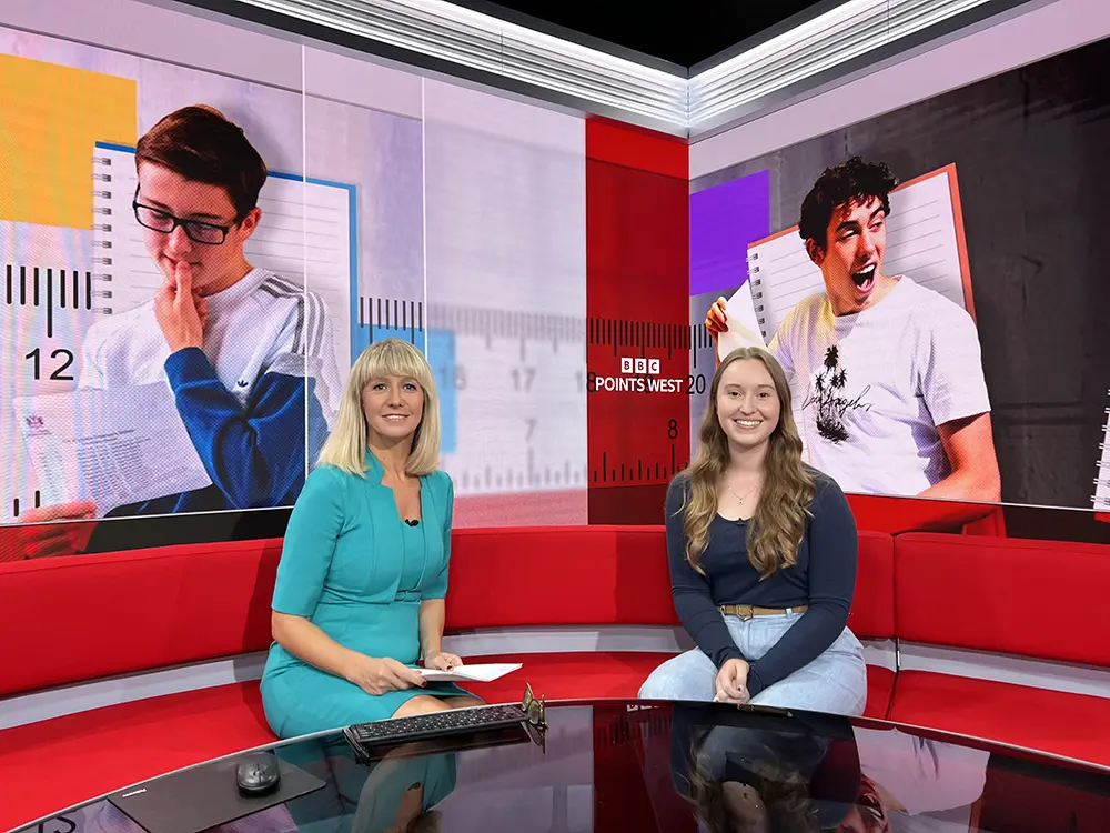 Two people seated on a red studio sofa during a BBC Points West broadcast, with large screens and graphics in the background.
