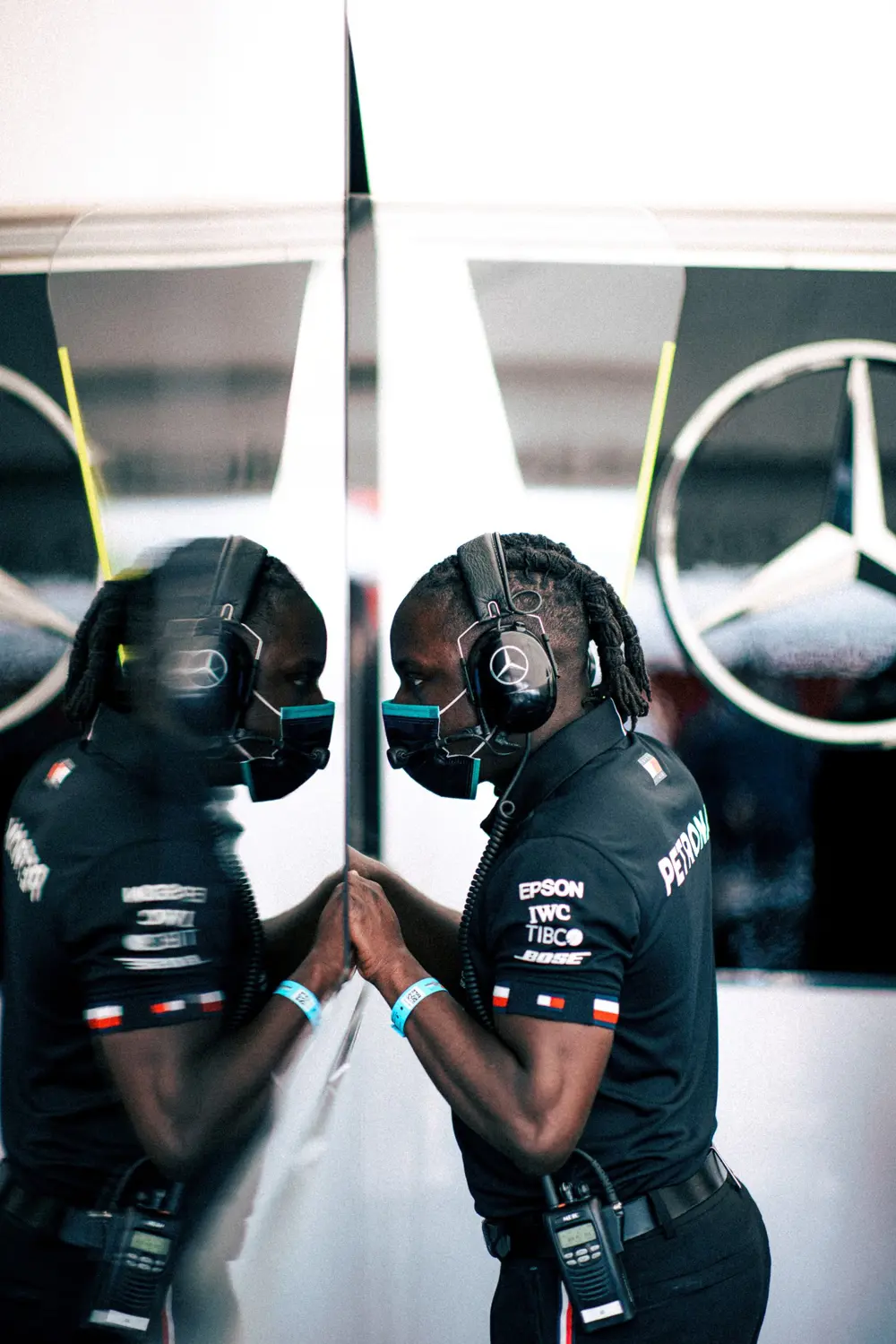 James Dornor, wearing a Mercedes engineer uniform, headset and face mask, stands facing his reflection, hands pressed against a glass wall in the garage.