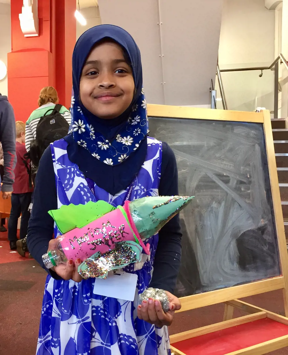 A child holding a rocket covered in glitter made out of a plastic bottle.