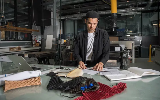 Ryan Mario Yasin in the studio behind a desk. He holds a ruler over his clothing designs and clothing samples can be seen on the desk in front of him. 