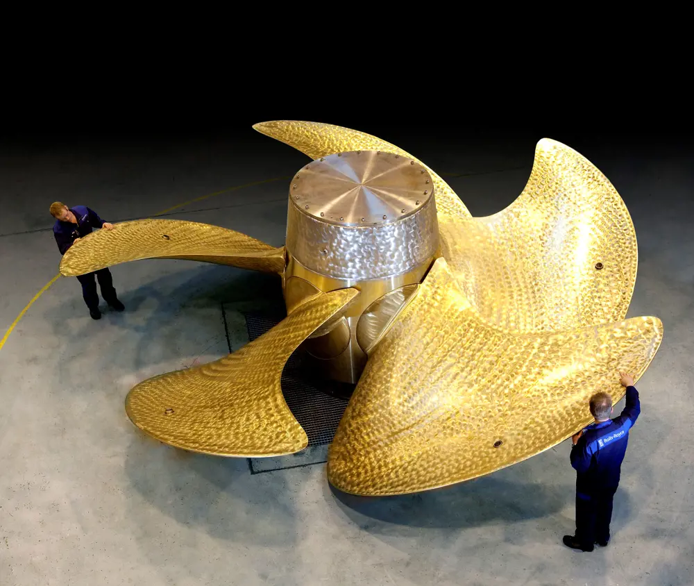 A large propeller on the floor with two people standing next to opposite blades.