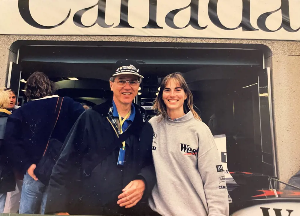 An older man in glasses and woman in her twenties grinning and wearing racing merch, standing in front of a racecar.