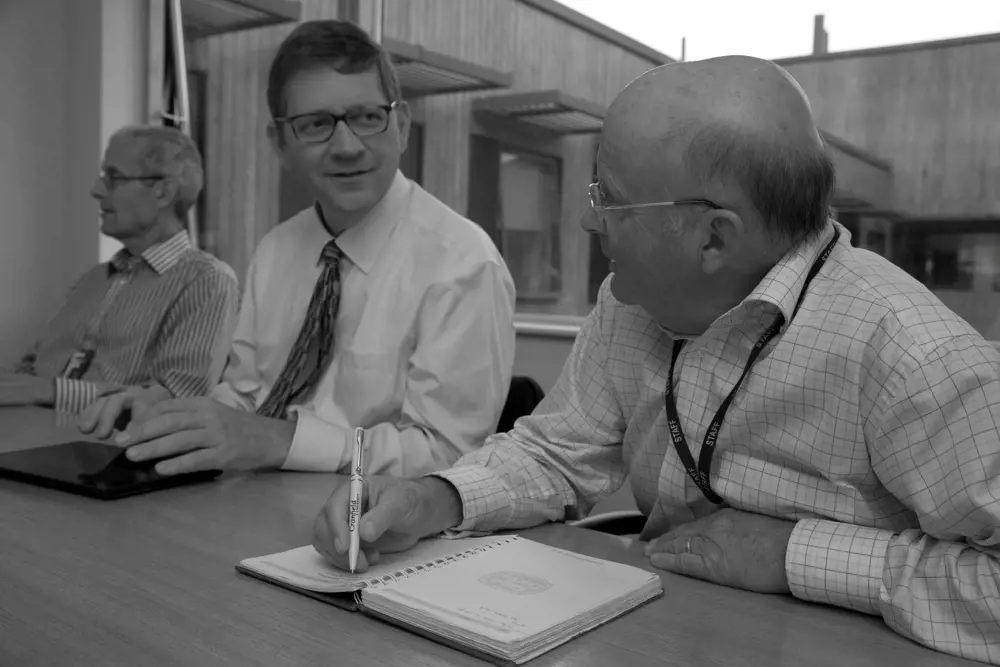 Black and white photograph of three men in business attire, sitting at a table in a meeting room with a pen and notebook.