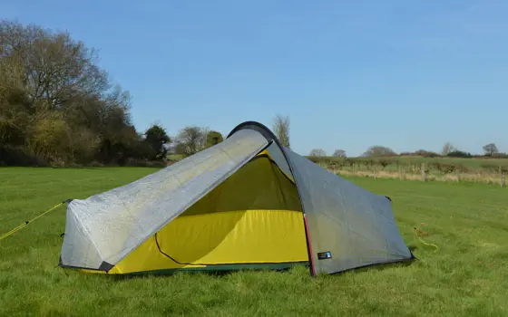 A terra nova tent in a field,  with a grey outside and yellow inside lining. It is held up by a semi-circular polar in the middle, and vertical pole at each end, with pegs to keep the fabric in place. 